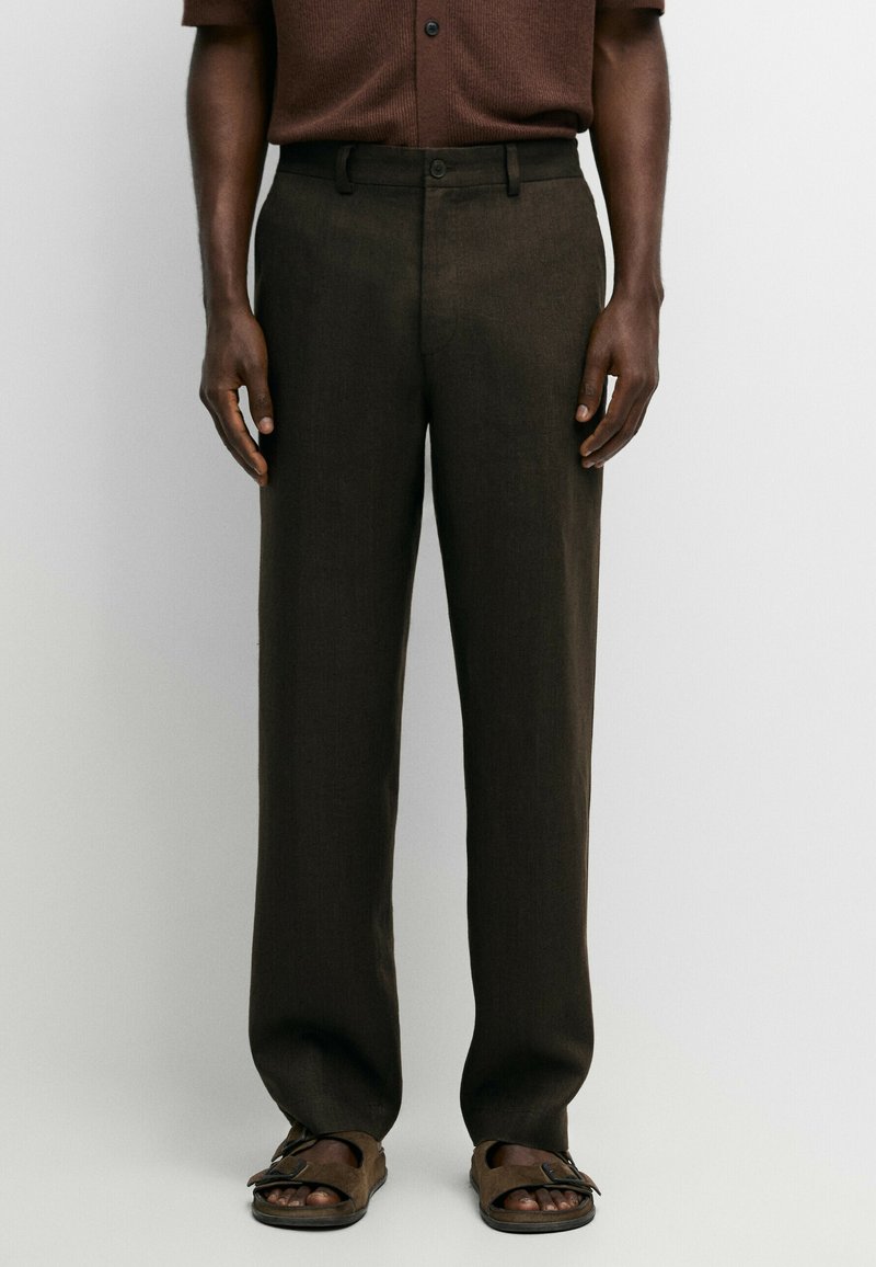 Man wearing dark brown straight-leg trousers paired with matching brown sandals and a brown short-sleeve shirt against a plain background.
