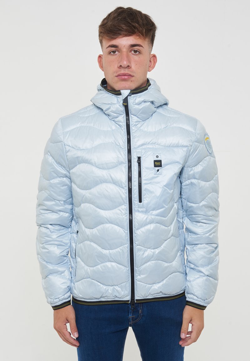 Giacca puffer azzurro chiaro con motivo a onde trapuntato, chiusura con zip, tasche frontali e cappuccio. Dettagli neri sulla zip e sui polsini.
