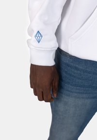 Felpa bianca con polsini a coste, caratterizzata da un logo a forma di diamante ricamato in azzurro chiaro sulla manica sinistra. Abbinata a jeans blu sbiaditi.