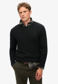 Zwarte gebreide trui over een groen en marineblauw geruit shirt, gecombineerd met olijfgroene broeken. Horloge om de pols, staand tegen een witte achtergrond.