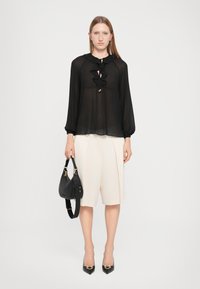 Black sheer blouse with ruffled neckline and long sleeves; cream-colored pleated shorts; black handbag; black pointed-toe heels.
