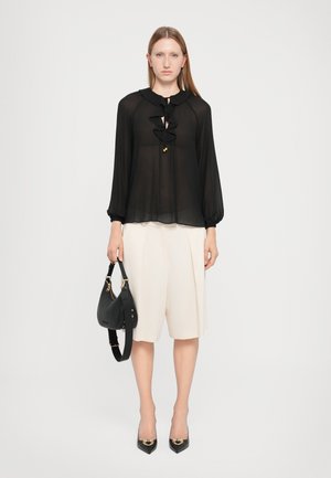 Blusa negra transparente con escote con volantes y mangas largas; pantalones cortos plisados de color crema; bolso negro; tacones negros de punta.