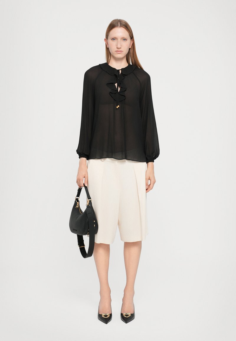 Black sheer blouse with ruffled neckline and long sleeves; cream-colored pleated shorts; black handbag; black pointed-toe heels.