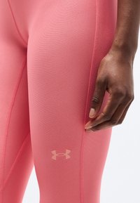 Κοντινό πλάνο του χεριού ενός ατόμου που ακουμπά δίπλα σε ροζ κολάν Under Armour με ορατές ραφές και λογότυπο στο μηρό.