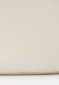 Borsa bianca in pelle con superficie texture e logo dorato stampato "FURLA" sul davanti. Bordature lisce e design minimalista.