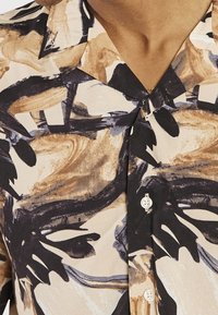 Lyhythihainen beige paita, jossa rohkeat mustat ja ruskeat abstraktit kuviot, lovikaulus ja valkoiset napit edessä. Sileä kangaspinta.