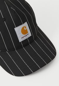Svart caps med hvite striper, med en firkantet hvit lapp som viser en gul logo. Laget av vevd stoff med en buet skjerm.