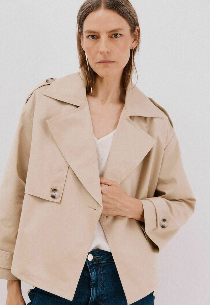 Beige kort jacka gjord av slät tyg, med stor krage, framknäppning och vida ärmar med knappdetaljer.