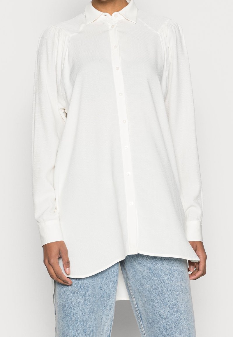 Camicia bianca con tessuto testurizzato, maniche lunghe e orlo curvo. Presenta un colletto classico e spalle leggermente imbottite.