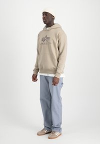 Sudadera beige con bolsillo frontal y logo, superpuesta a una camisa blanca. Pantalones azul claro y zapatillas grises con acentos blancos completan el atuendo.