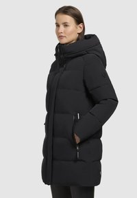 Fekete puffermellény kapucnival, steppelt designnal, elülső cipzárral és oldalsó cipzáras zsebekkel. Puha tapintású, térdközépig ér.