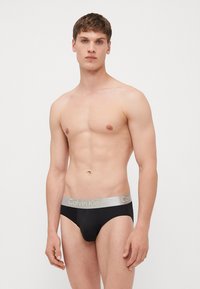 Schwarze Unterhose mit einem silbernen Bund, der "Calvin Klein" in goldenen Buchstaben trägt. Glattes Material, körperbetonter Schnitt, minimalistisch im Stil.