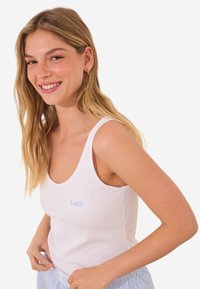 Bílý žebrovaný top na ramínka s širokými pásky a světle modrým vyšitým textem "Lazy." Model má na sobě světle modré pruhované kalhoty, které ukazují uvolněný střih.
