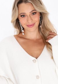 Cardigan alb cu un design în formă de V, butoane decorative mari și textură moale; accesorizat cu cercei lungi, cu accente de perlă.