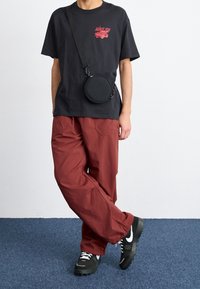 Svart bomulls-T-shirt med röd grafik, löst sittande röda byxor och svarta sneakers med vita detaljer. Inkluderar en rund svart axelväska.