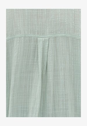 Hellgrüne, strukturierte Stoffe mit einem dezenten Karomuster und einem zentralen Faltendetail auf der Rückseite, was einen weichen Fall erzeugt.