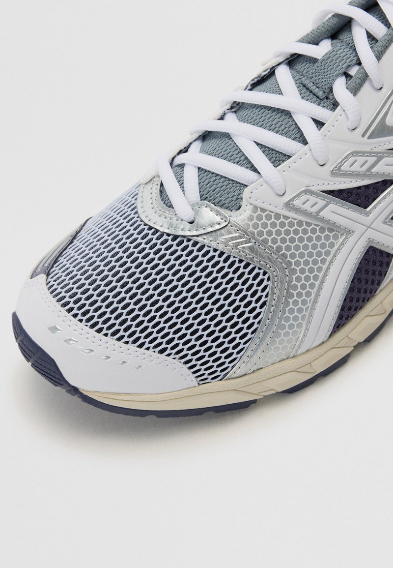 Zapatilla deportiva con parte superior de malla en gris claro y azul marino con detalles en plata, puntera redondeada y cordones blancos. Suela texturizada para tracción.