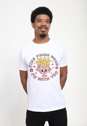 Hombre sonriente con camiseta blanca que tiene un dibujo de papas fritas sonrientes, corazones y el texto: "Time fries when I'm with you,", posando sobre un fondo liso.