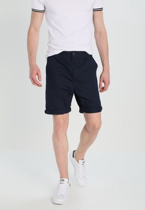 Marineblå bomulds shorts med opkrøllede kanter, med en frontknaplukning og lommer. Båret med en hvid kortærmet skjorte og hvide sneakers.