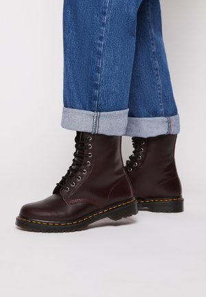 Botas de tornozelo em couro bordô com atacadores pretos, ponta arredondada, costura amarela e sola de borracha grossa, combinadas com jeans azuis com as bainhas viradas.
