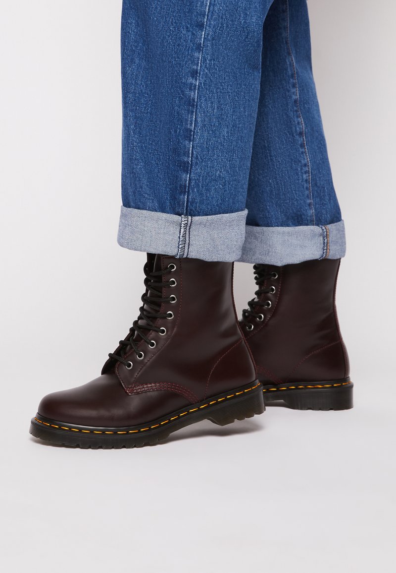 Botas de tornozelo em couro bordô com atacadores pretos, ponta arredondada, costura amarela e sola de borracha grossa, combinadas com jeans azuis com as bainhas viradas.