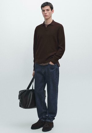 Giovane in piedi che indossa un maglione a costine marrone scuro, jeans blu, scarpe scure, tenendo una borsa da viaggio in pelle nera al suo fianco.