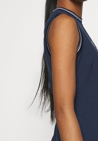 Side view of a person with long dark hair wearing a sleeveless navy blue top with white striped trim on the armhole and neckline.