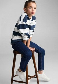 Pruhovaná námořnická modrá a bílá mikina, tmavě modré džíny, bílé tenisky. Dítě sedící na dřevěné stoličce, ruce položené na kolenou.