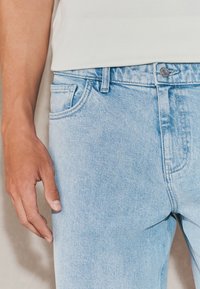 Light blue denim jeans with a slim fit, featuring a front pocket, metal button closure, and minimalistic stitching details.