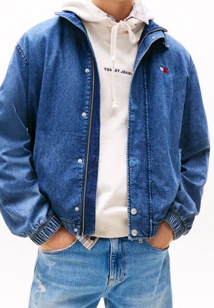 Mann trägt blaue Jeansjacke über weißem Tommy Jeans Hoodie und hellblaue, abgenutzte Jeans, Hände in den Taschen, steht vor weißem Hintergrund.