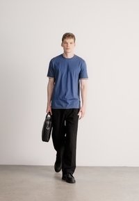 Kék rövid ujjú póló kontrasztos szegéllyel, fekete nadrággal és fényes fekete cipőkkel párosítva, fekete bőrtáskát tartva.