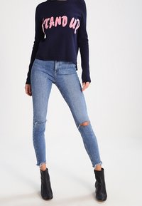 Marinblå tröja med rosa "STAND UP"-text, tillsammans med ljusblå rivna skinny jeans och svarta ankle boots. Avslappnad outfit, mjuk textur.