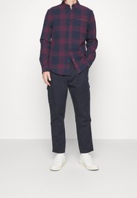 Camicia a maniche lunghe a quadri in blu navy e bordeaux, con colletto all'italiana e tasche decorate; abbinata a pantaloni cargo scuri e sneaker bianche.