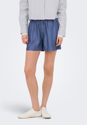 Model iført blå denimshorts med snøre, stribet langærmet skjorte og hvide sneakers, stående på en lys baggrund.