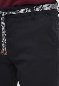 Black trousers made of cotton with a textured white and black patterned belt. Features two side pockets and brown leather accents.