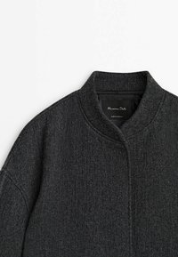 Veste gris foncé avec un col rond, une silhouette structurée, un tissu texturé et un design minimaliste avec des détails de couture subtils.