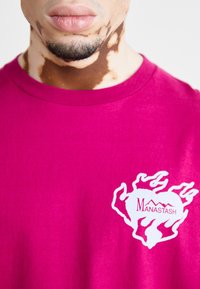 Fuksjowa bawełniana koszulka z okrągłym kołnierzykiem i krótkimi rękawami, zdobiona białym logo w kształcie płomienia z napisem "Manastash" na lewej stronie klatki piersiowej.