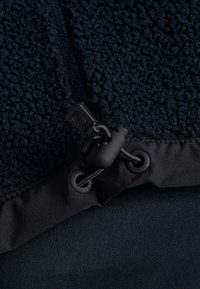 Tissu en polaire bleu foncé avec une surface texturée, comportant un cordon de serrage et un tissu lisse le long du bord. Détails noirs sur le matériel.