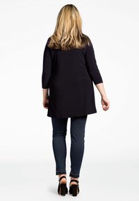 Zwarte tuniek met driekwart mouwen, gecombineerd met donkere skinny jeans en zwarte hoge hakken, gezien vanuit de achterkant.