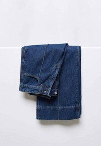 Jean en denim bleu plié en deux, suspendu à une barre horizontale blanche contre un mur blanc uni.