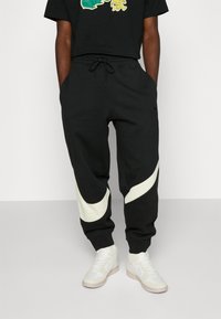 Czarne dresy z kremowym, zakrzywionym paskiem, posiadające elastyczny pas ze sznurkiem oraz boczne kieszenie. Noszone z białymi trampkami.