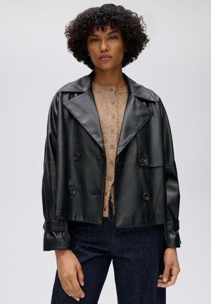 Femme aux cheveux courts et bouclés portant une veste noire en cuir courte et croisée sur un pull beige boutonné et un jean foncé.