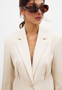 Blazer beige avec des revers marqués et une fermeture à un bouton, associé à des lunettes de soleil surdimensionnées en écaille de tortue et des cheveux lissés en une queue de cheval basse.