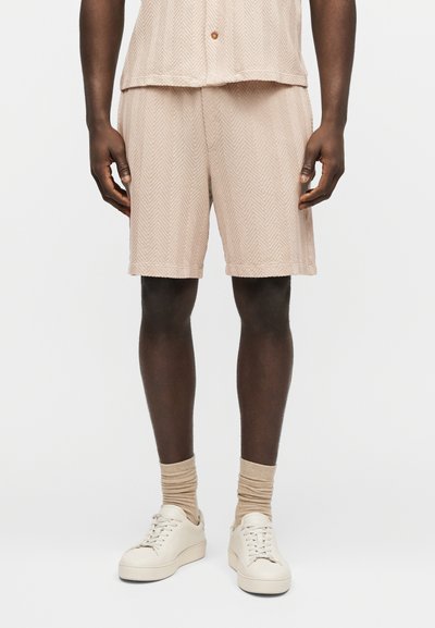 Personne portant un short beige texturé, une chemise assortie, des chaussettes beiges et des baskets blanches à lacets sur un fond uni.