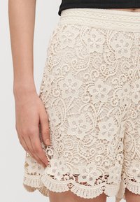 Lace shorts in ivory with floral patterns, scalloped hem, and a textured waistband. Features intricate stitching and open weave design.