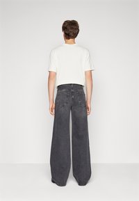Jeune personne de dos, portant une chemise blanche à manches courtes et un pantalon en jean large taille haute gris foncé, devant un fond uni.