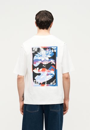 Személy fehér pólót visel, amelyen színes, absztrakt virágmintázat és P, U, M, A betűk találhatók a hátoldalon, kék farmernadrággal párosítva.