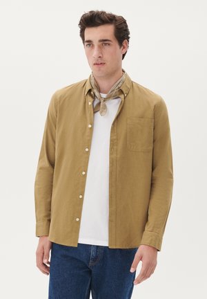 Khaki button-up shirt with a single chest pocket, white T-shirt underneath, paired with blue denim jeans and a patterned beige scarf.