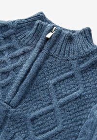 Pull en laine bleu tricoté avec un col montant côtelé et une demi-fermeture éclair argentée, présentant des motifs texturés en losanges et torsades.