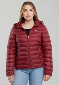 Žena nosící bordó prošívanou péřovou bundu s kapucí a zipem, v kombinaci se světle modrými džínami, stojící před jednobarevným pozadím.